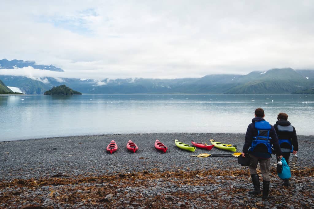 Two people walk toward a row of kayaks on a rocky beach